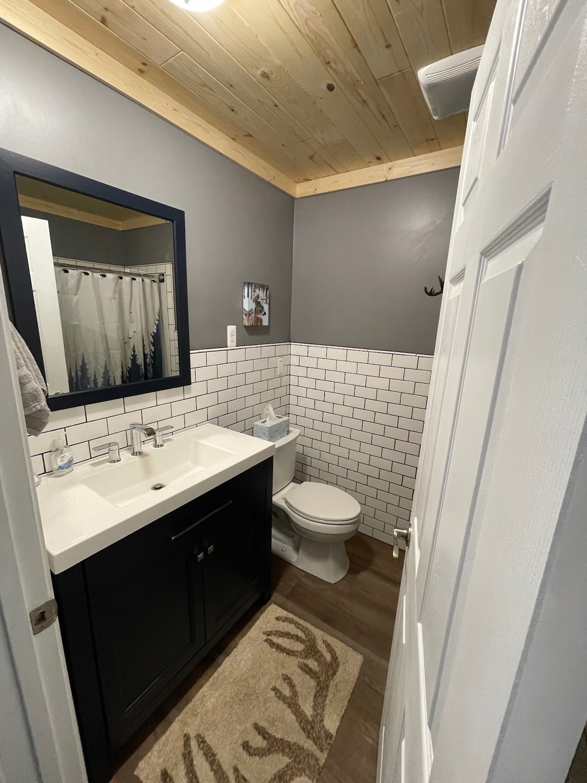 A modern bathroom features a wall-mounted mirror above a dark cabinet with a double sink. A white tiled wall complements the wood-paneled ceiling. A beige area rug with an antler design lies on the floor, while a patterned shower curtain adds a touch of privacy.