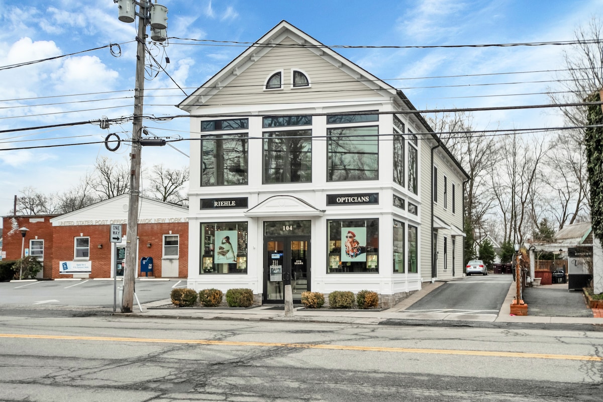 An attractive two-story building with large windows showcases a modern façade. A clear sign indicating 'Opticians' is displayed prominently. The surrounding area includes a parking lot and well-maintained landscaping, creating an inviting atmosphere. Power lines are visible, set against a blue sky.