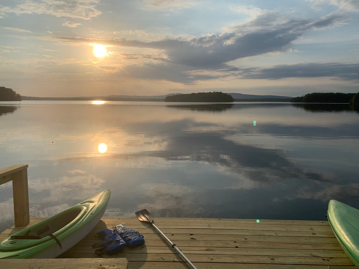 A serene view of the lake is captured with the sun setting on the horizon, reflecting shimmering colors on the calm water. A wooden dock is adorned with two kayaks and an oar, inviting an evening adventure on the lake.