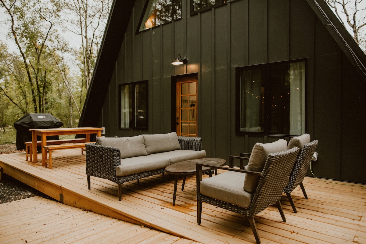 A spacious deck features outdoor seating with a plush couch and two armchairs surrounding a coffee table. A dining table is positioned nearby, complemented by a barbecue grill. Large windows in the back of the cottage provide views of the surrounding forest.