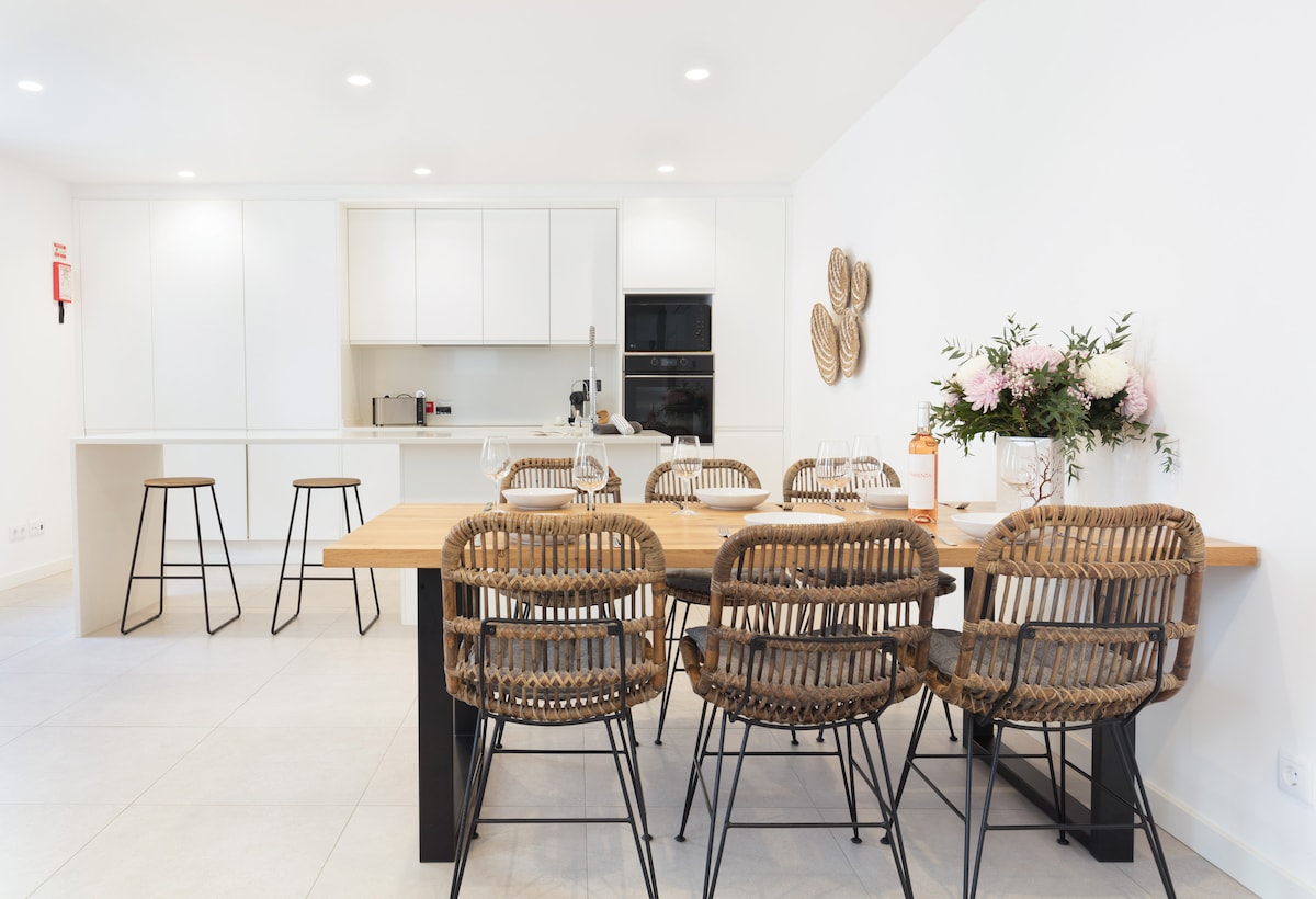 A modern kitchen and dining area features a spacious wooden table surrounded by six woven chairs. The sleek cabinetry and appliances create a streamlined appearance, while soft lighting enhances the inviting space. A floral arrangement decorates the table, adding a touch of elegance.