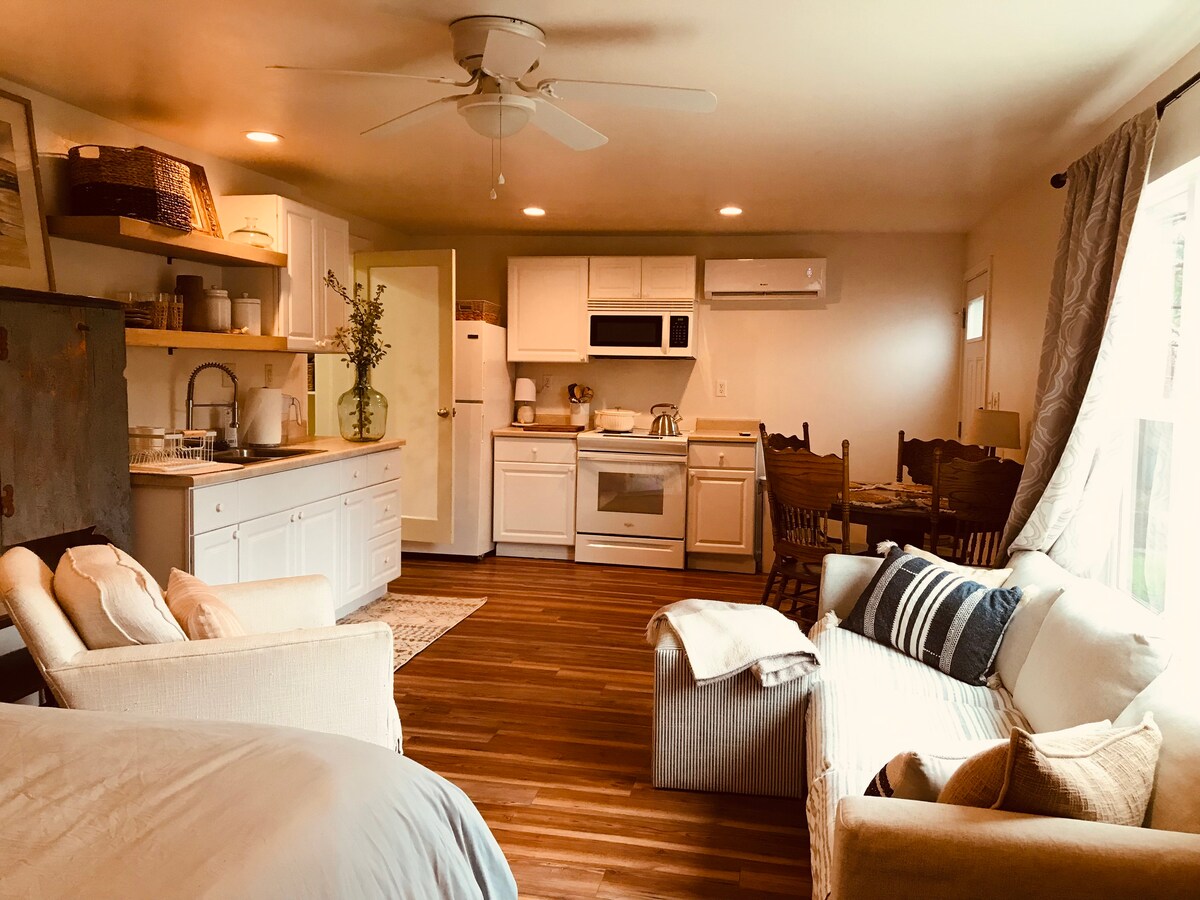 A cozy open-concept living space showcases a fully equipped kitchen with white cabinetry and modern appliances. Natural wood flooring complements a dining area with a small table and chairs. Comfortable seating is provided by a sofa and armchair, with light filtering through the large windows.