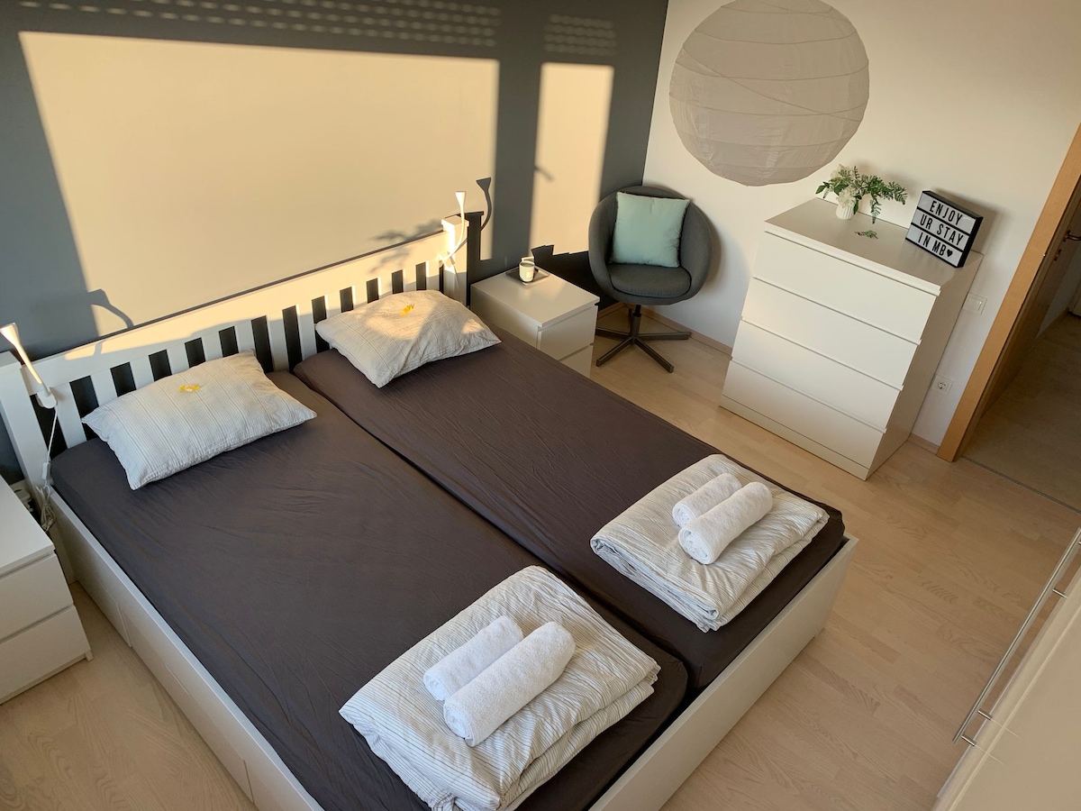 The sleeping area features a spacious double bed with dark bedding and two neatly arranged towels at the foot. A light-colored chest of drawers stands nearby, accompanied by a comfortable chair. Natural light filters through the room, creating a calm and bright environment.