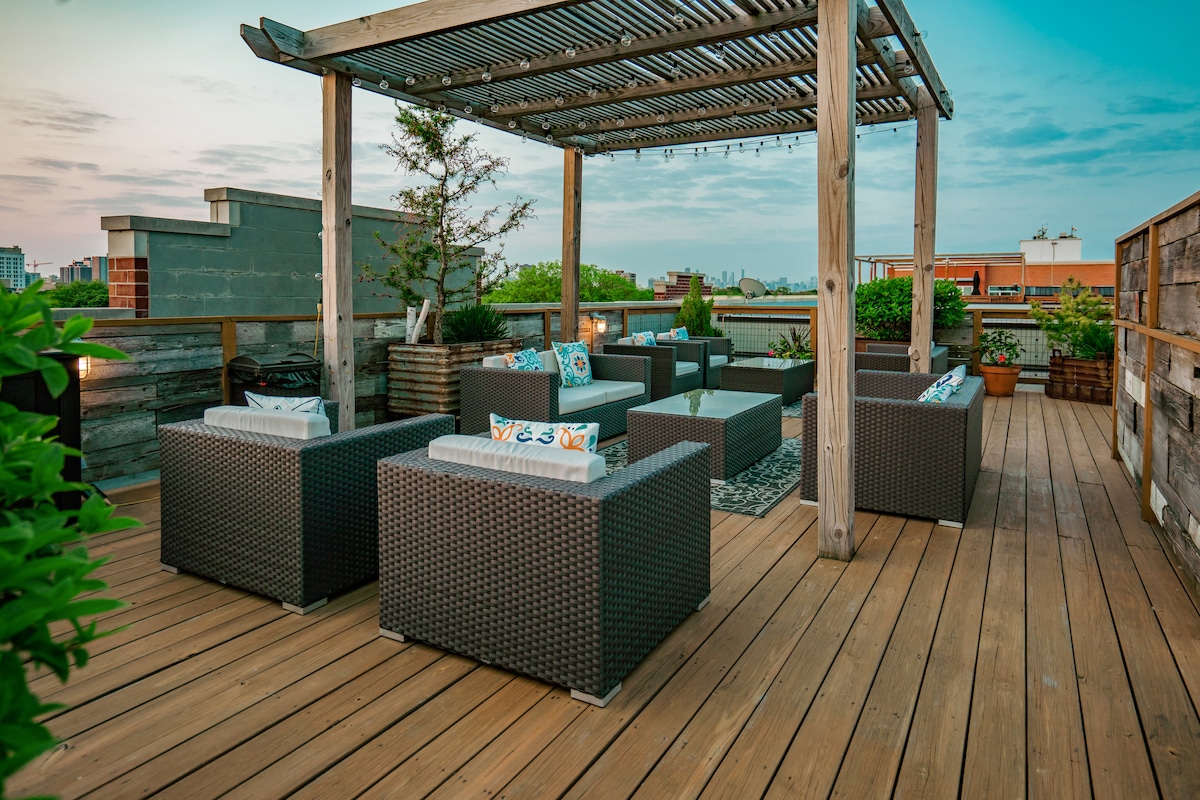 An outdoor rooftop space features comfortable seating arranged in a conversational layout, surrounded by greenery. A wooden pergola provides shade over the area, while decorative cushions add color. The deck's wooden flooring complements the serene setting, creating an inviting space for relaxation.