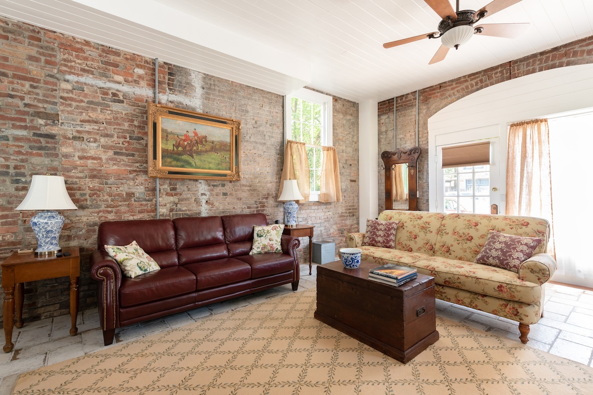 A welcoming living area features exposed brick walls, highlighting a vintage painting. A leather sofa and a floral upholstered couch are positioned around a wooden coffee table. Natural light enters through tall windows, complemented by sheer curtains that gently frame the view.