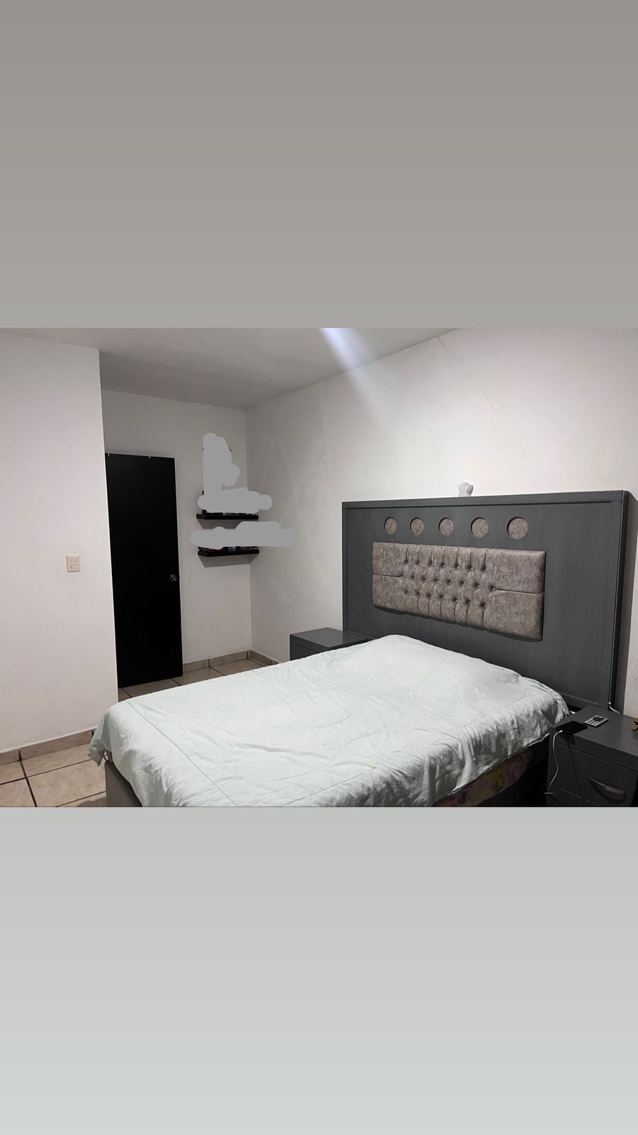 A spacious bedroom features a tufted headboard with circular accents and a light-colored blanket draped over the bed. A dark cabinet sits beside the bed, and a door leads to another room. Additional wall shelves are visible in the corner.