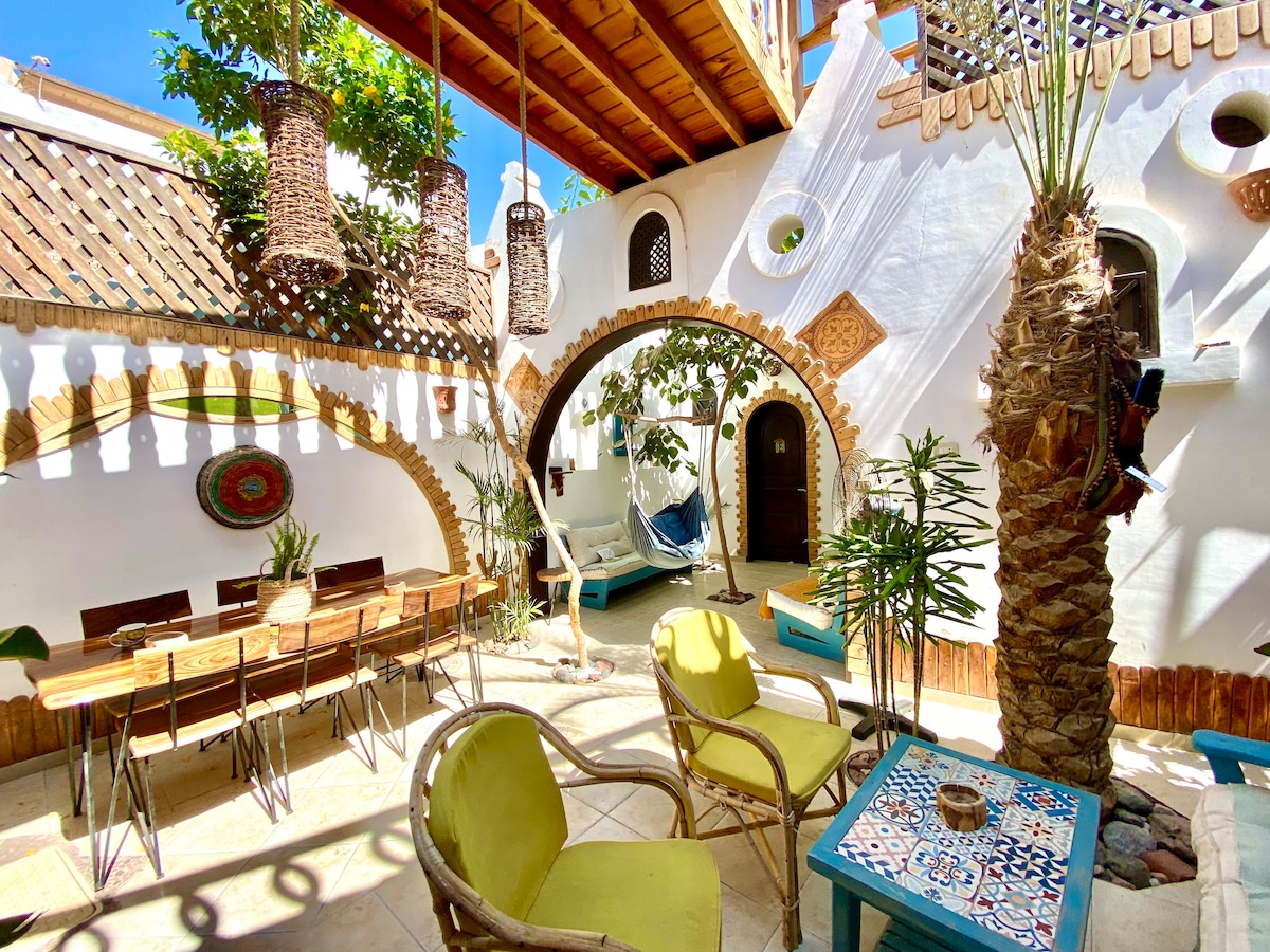 A vibrant outdoor space features a large dining table surrounded by colorful chairs. Lush greenery and hanging planters create a natural ambiance. Arched doorways and textured walls contribute to the Moroccan design, while a hammock offers a cozy spot for relaxation.