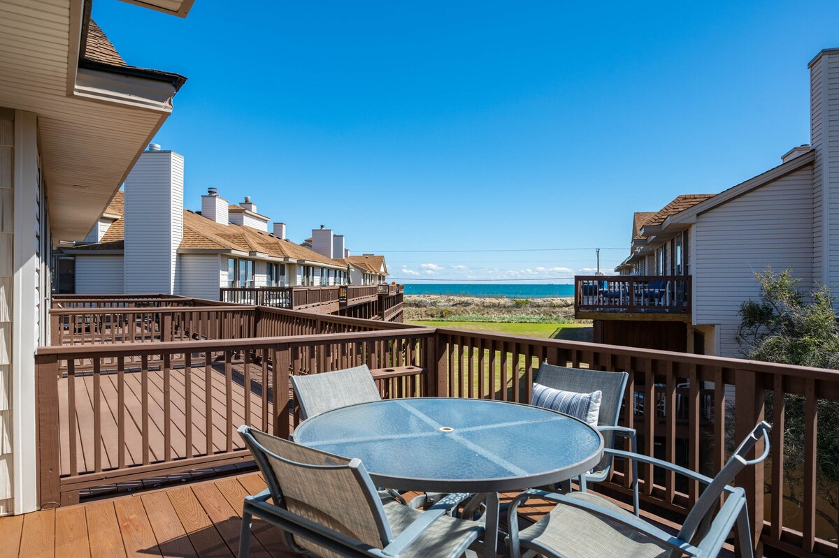 A deck area is presented with a round table and several chairs, surrounded by wooden railings. The view extends to the ocean, with a clear blue sky overhead and grass in the foreground. Nearby townhouses are visible in the background, enhancing the coastal setting.