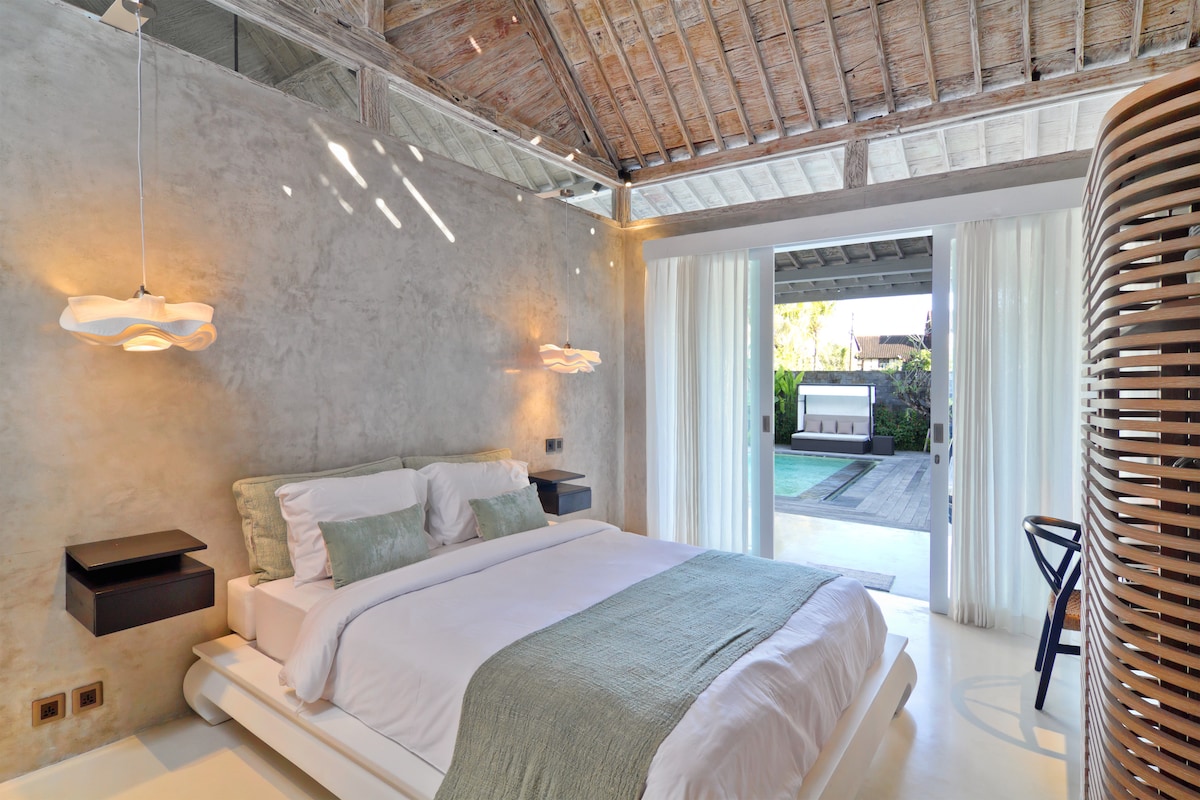A bright bedroom features a spacious bed with light bedding and textured pillows. Large glass doors open to an outdoor space, allowing natural light to fill the room. Textured walls complement the modern decor, while a unique wooden screen adds visual interest.