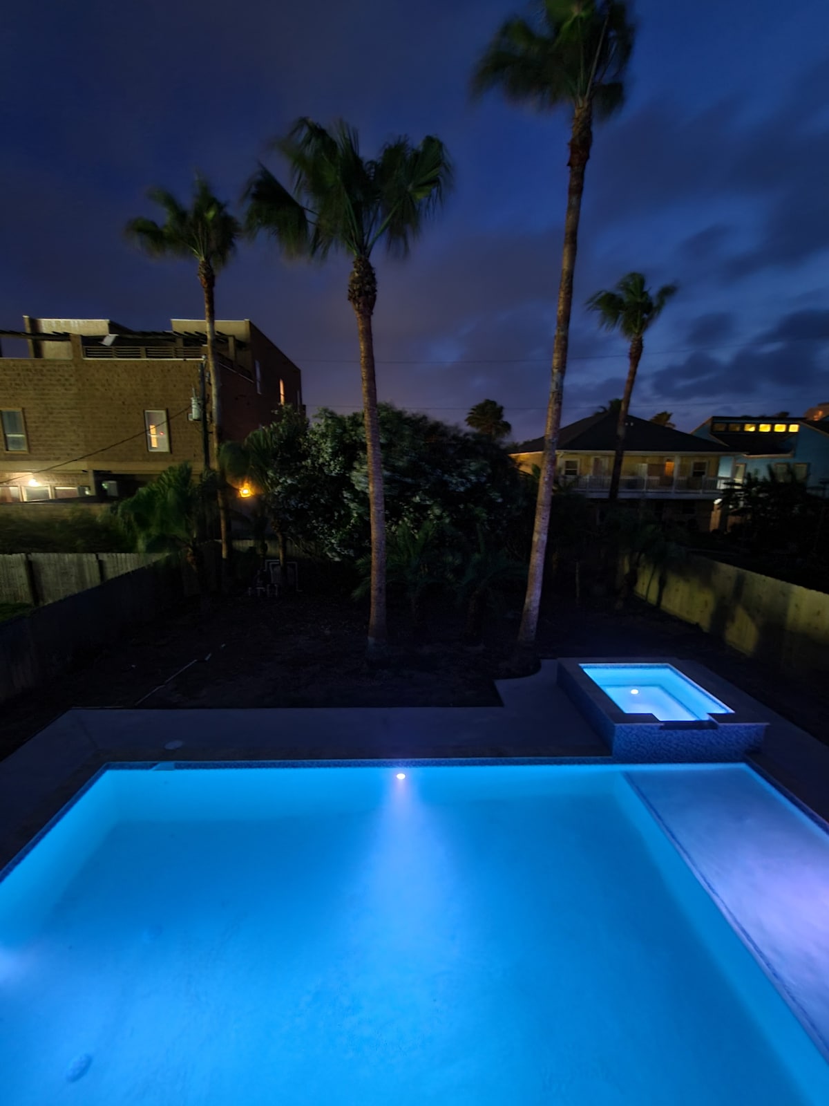 A spacious pool and hot tub are illuminated in calming blue tones, surrounded by multiple palm trees. The night sky casts a gentle glow over the backyard, emphasizing the tranquil atmosphere of the outdoor space.