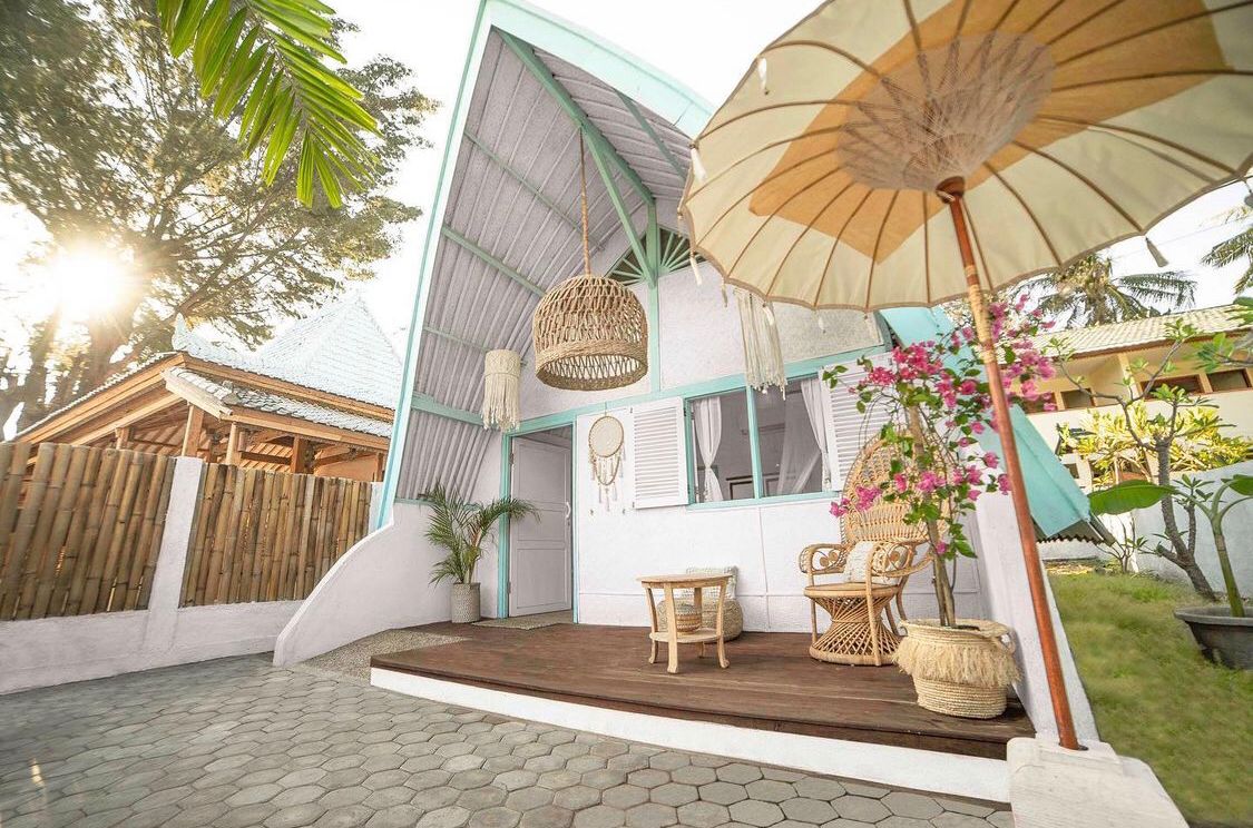 A charming cottage features an A-frame design with pale blue accents. A wooden deck furnished with a small table and chairs is shaded by a decorative umbrella. Tropical plants frame the entrance, and open shutters allow natural light to fill the space.