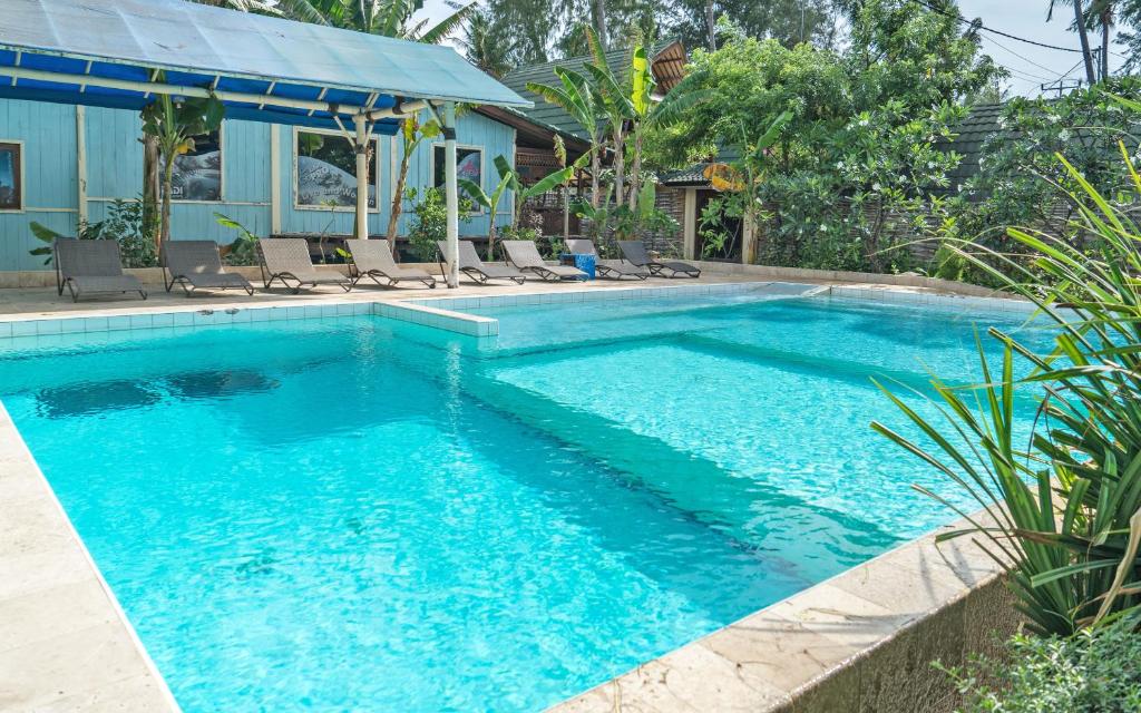 A clear swimming pool is surrounded by lounge chairs and lush tropical greenery. Sunlight reflects off the water, creating a serene atmosphere. A covered area features additional seating, providing a shaded retreat near the pool.