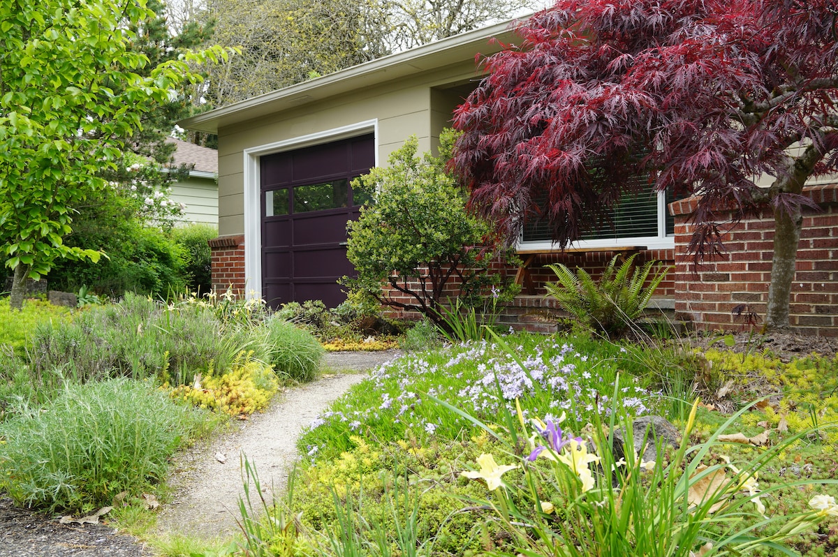 The exterior garden features a winding pathway surrounded by a variety of plants, including lush greenery and colorful flowers. A maple tree with red foliage stands prominently, alongside a garage door in a deep hue, all contributing to a charming and inviting entrance.