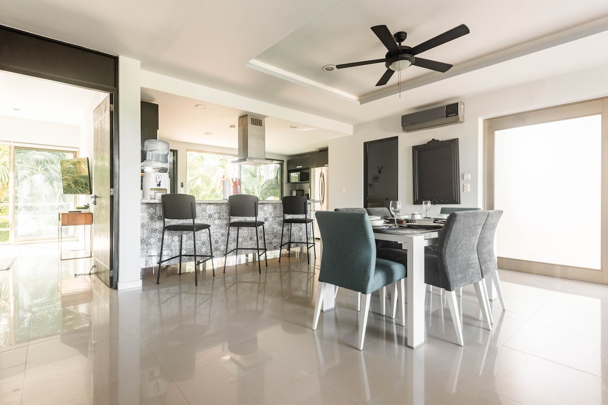 A spacious dining area features a table with seating for six and a ceiling fan. The open layout connects to a modern kitchen with bar seating. Large windows allow natural light to brighten the space, while views of greenery are visible in the background.