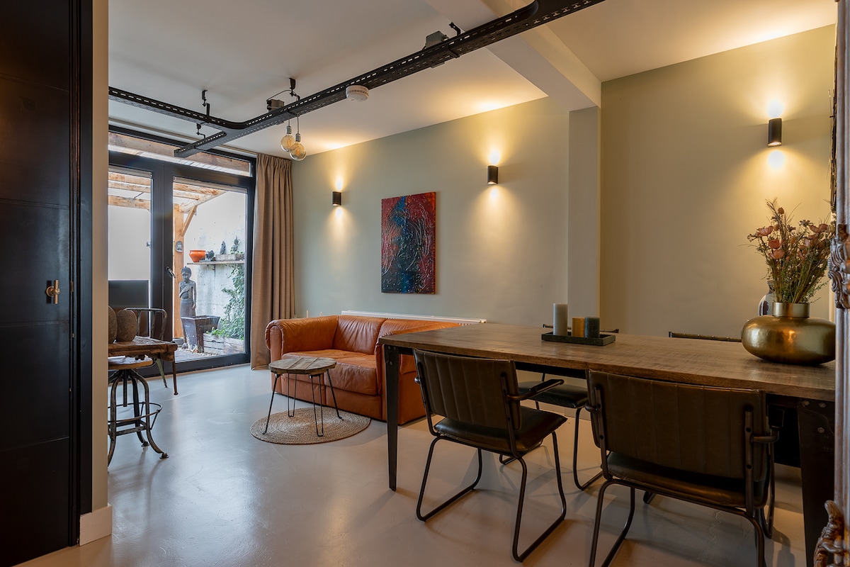 A modern living space is illuminated by warm wall lights. A spacious leather sofa is positioned near a dining table with metal chairs. An art piece adorns the wall, and natural light enters through large doors that lead to the outdoor area.