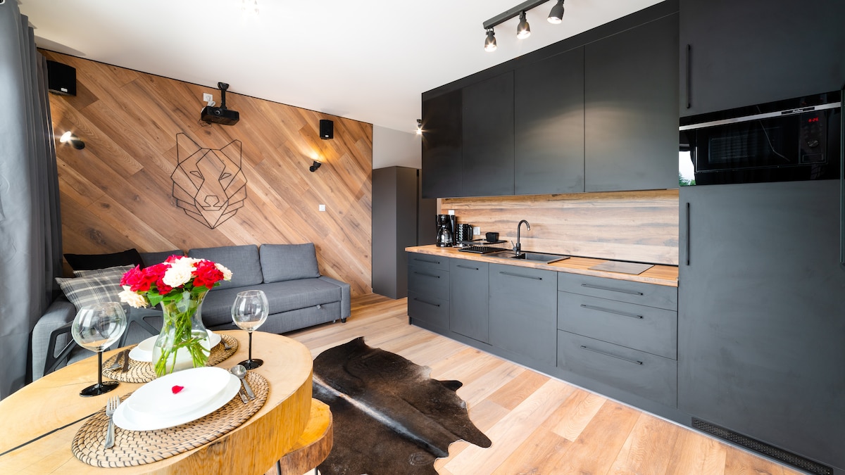 The studio apartment features an open kitchen with modern appliances and wooden countertops. A cozy seating area includes a gray sofa against a wood-paneled wall adorned with a decorative animal motif. A dining table is elegantly set with tableware and fresh flowers.
