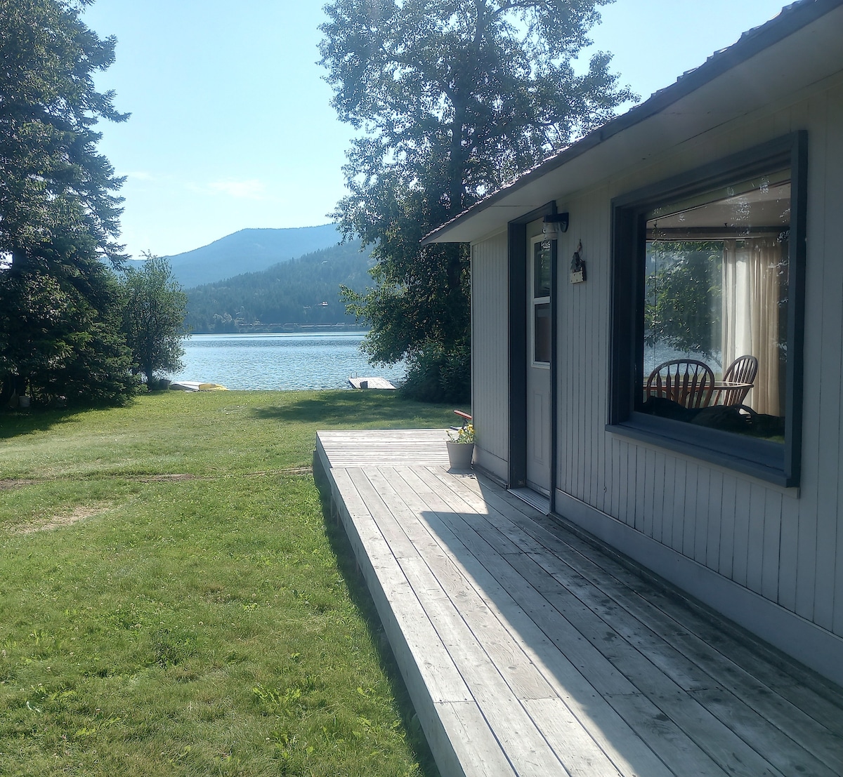 A serene view is captured from a wooden deck, showcasing a cabin adjacent to a calm lake. Lush greenery surrounds the area, complemented by distant mountains under a clear blue sky. Large windows invite natural light into the cabin, enhancing the tranquil setting.