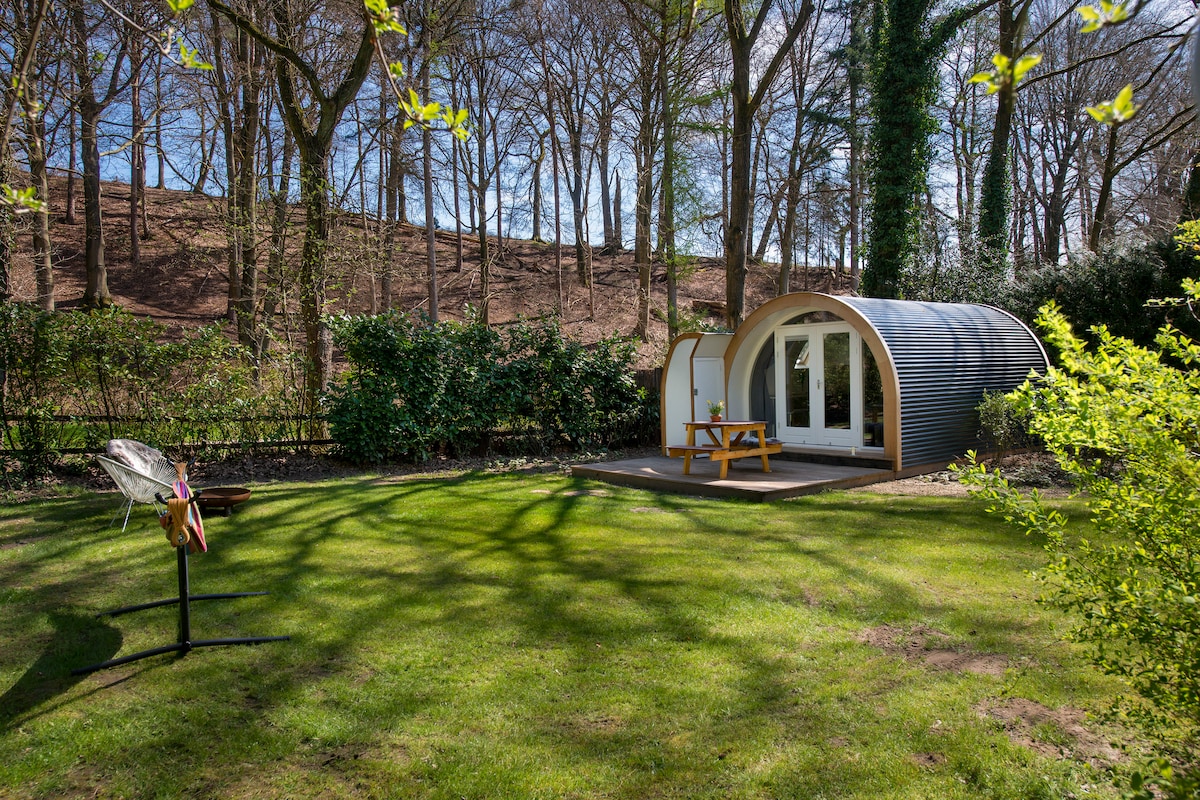 A unique accommodation is nestled in a serene green garden, surrounded by trees. The structure features a curved design and large glass doors, allowing natural light to enter. A wooden picnic table sits on a deck outside, complemented by a grilling station nearby.