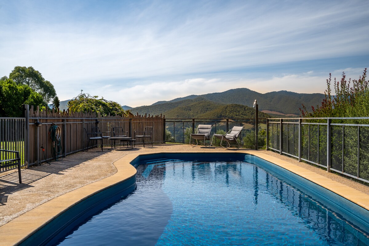 The inground swimming pool is framed by a sun deck and lounge chairs, offering scenic views of the surrounding hills and valleys. Lush greenery can be seen along the property, creating a serene outdoor space for relaxation.