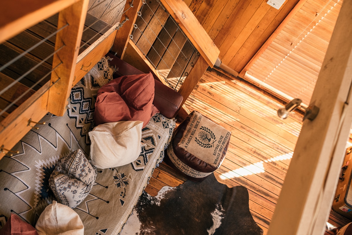 An overhead view of a cozy mezzanine area features various cushions scattered on a textured rug. Natural light filters through wooden blinds, illuminating the warm timber flooring. The space includes a mix of seating options, creating an inviting atmosphere for relaxation.