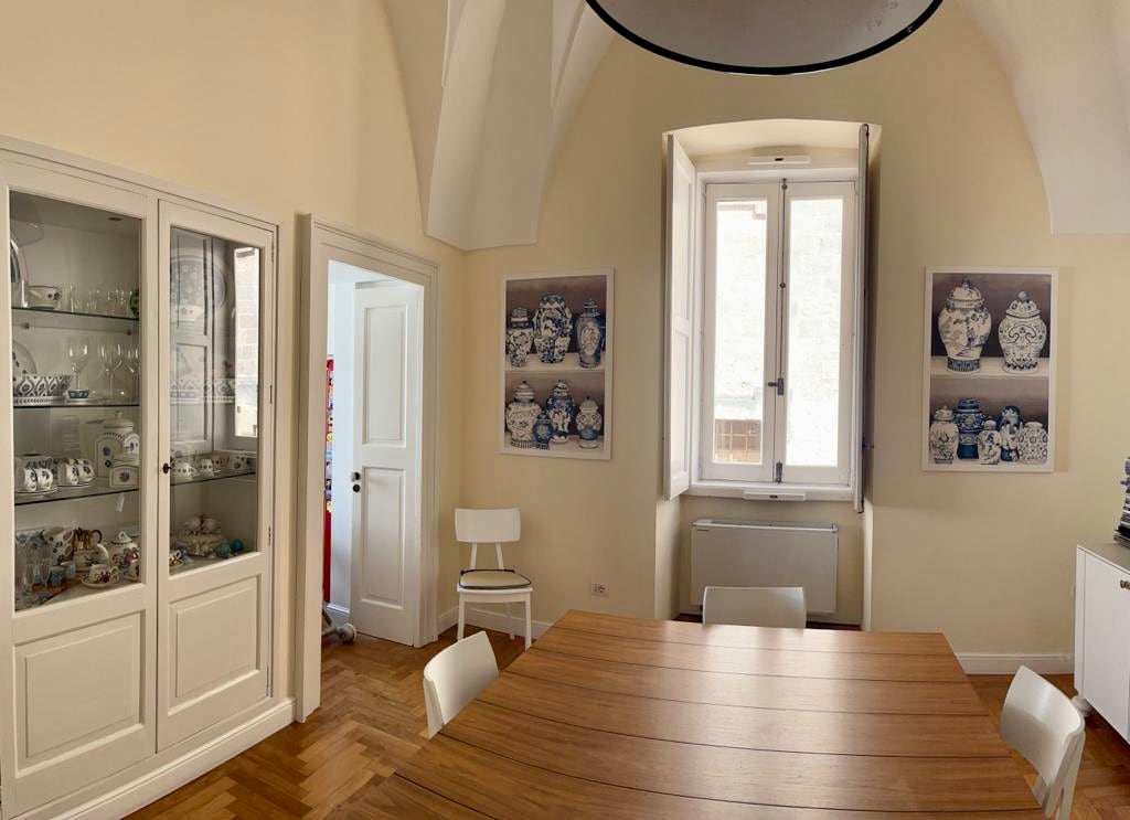A dining area showcases a wooden table surrounded by white chairs. Large windows allow natural light to fill the space, which is adorned with framed art featuring decorative ceramic pieces. A glass cabinet displays an elegant collection of dishes, enhancing the room's bright and airy feel.