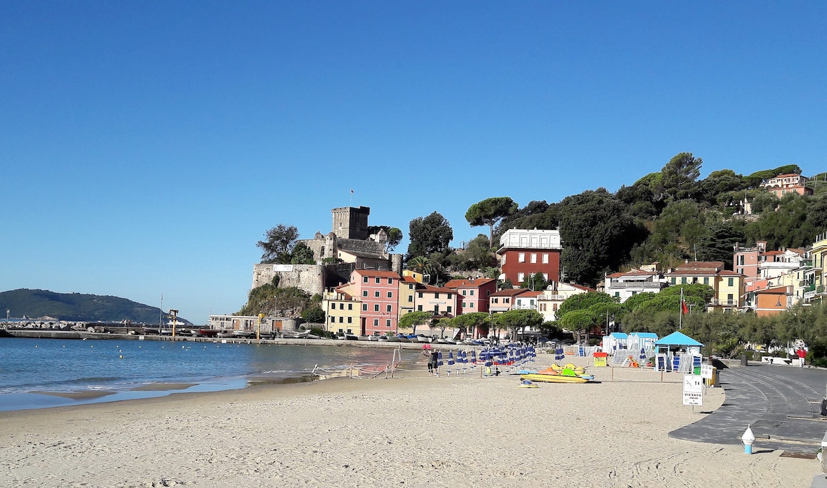 A sandy beach is shown, with gentle waves lapping at the shore. Colorful beach umbrellas are arranged in neat rows. In the background, a medieval castle and a cluster of charming buildings are visible, surrounded by lush greenery and clear blue skies.