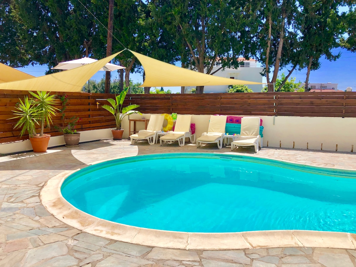 A inviting outdoor pool area is surrounded by stone flooring and features comfortable loungers. Sunshades provide relief from the sun, while potted plants add a touch of greenery. A wooden fence offers privacy, creating a serene setting for relaxation.