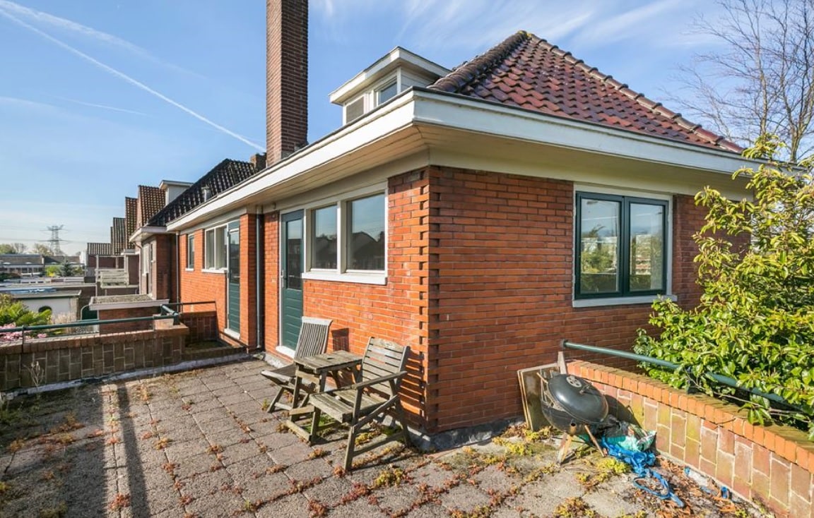 An outdoor terrace is shown, featuring several wooden chairs and a small table. Light-colored paving stones cover the ground, while greenery is visible along the edges. The brick wall of the building provides a warm contrast to the blue sky overhead.
