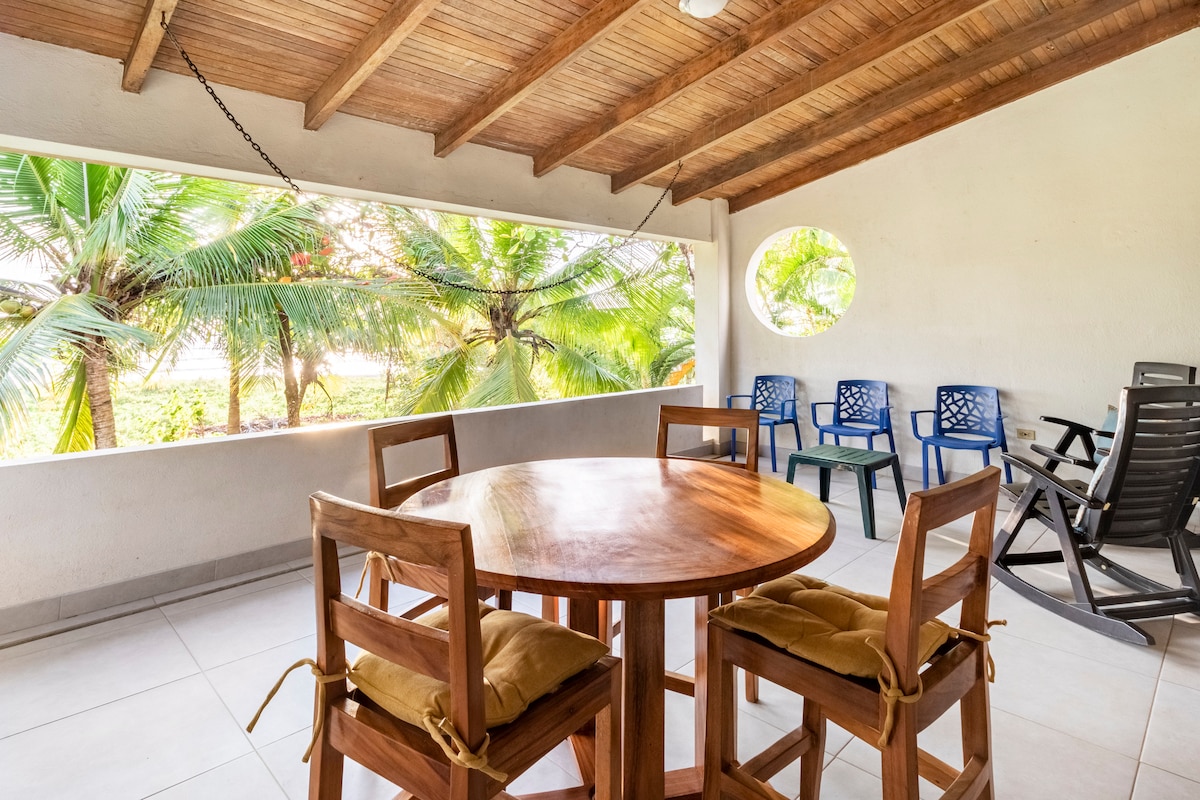 A covered outdoor terrace features a round wooden table surrounded by four high-backed chairs with cushions. Two additional chairs in the background provide seating, while large windows frame a view of lush palm trees, allowing natural light to fill the space.