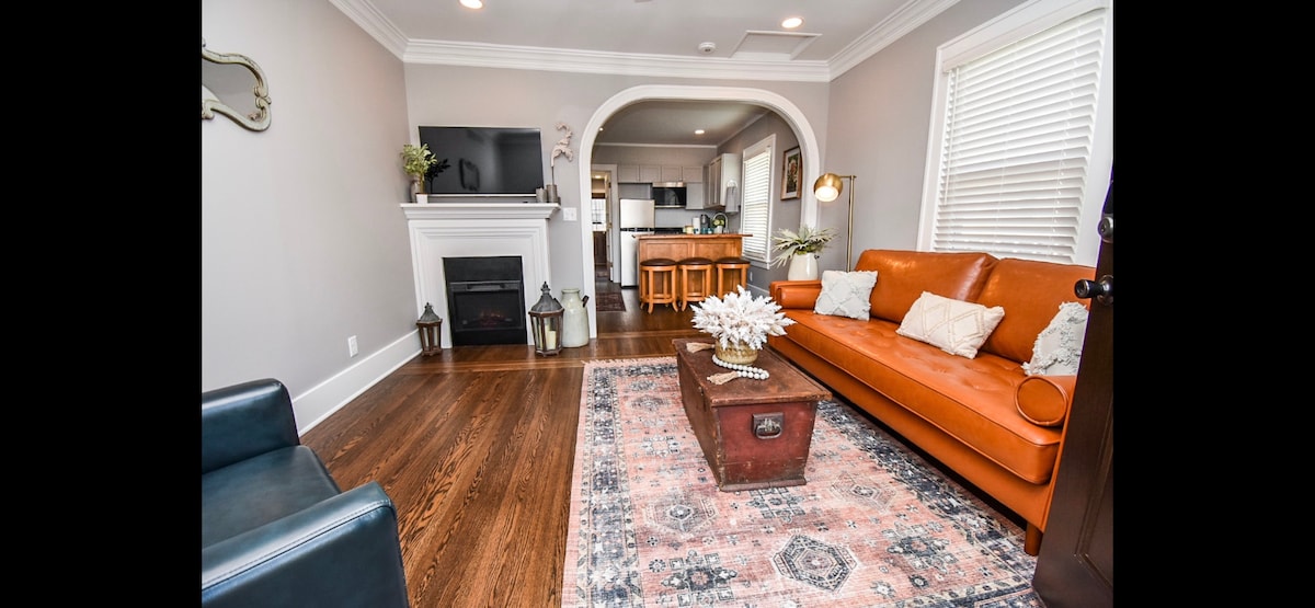 A warm living room showcases an orange sofa placed across from a stylish loveseat. A dark wood coffee table sits atop a patterned area rug. A fireplace is positioned beneath a wall-mounted TV. The room features natural light streaming through nearby windows.