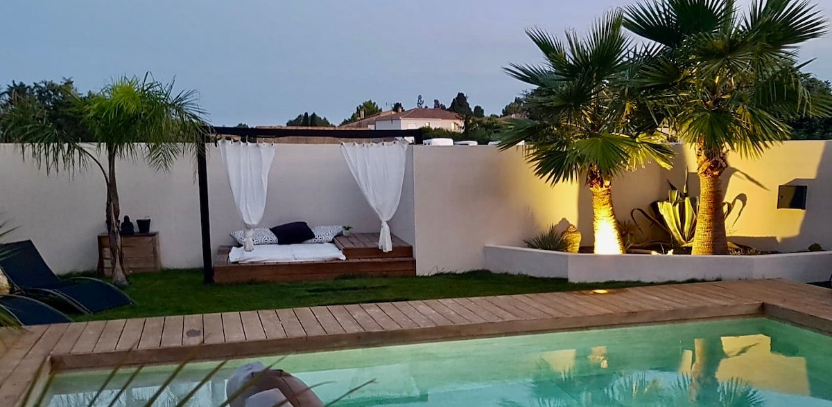 An outdoor area features a serene pool surrounded by wooden decking. A daybed with draped fabric is positioned beside the pool, complemented by landscaped greenery, including palm trees. Soft lighting enhances the tranquil atmosphere, creating an inviting space for relaxation.