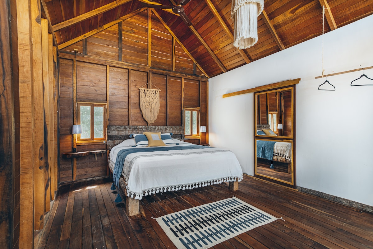 A spacious room features warm wooden walls and ceiling beams, complemented by a high ceiling. The comfortable bed is adorned with a light blanket and decorative pillows. Natural light enters through two windows, while a large mirror and hanging hooks are visible across the room.