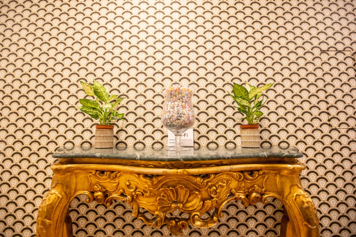 An ornate wooden console table is displayed against a textured patterned wall. Two potted plants with green leaves are positioned at either side of a decorative vase at the center, which is filled with colorful elements, adding a touch of elegance to the space.