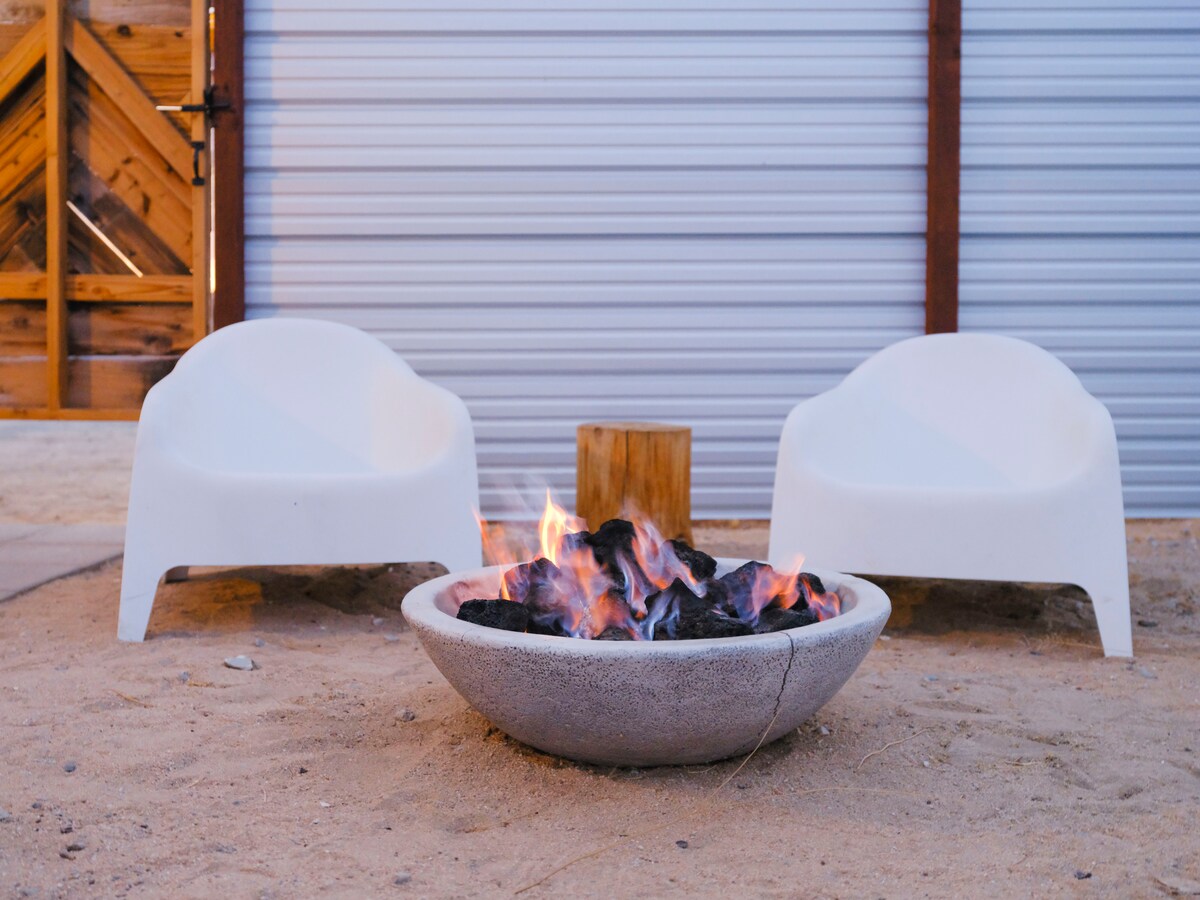 A fire pit surrounded by sand features glowing flames and a circular stone structure. Two white chairs are positioned nearby, offering a space for relaxation. A wooden stump serves as a natural side table, enhancing the outdoor setting.