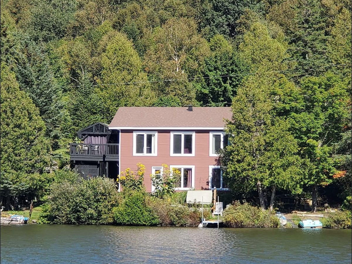 The house is set against a backdrop of lush trees, visible from across the lake. Large windows allow natural light to illuminate the exterior. A small dock extends into the water, providing access for recreational activities.