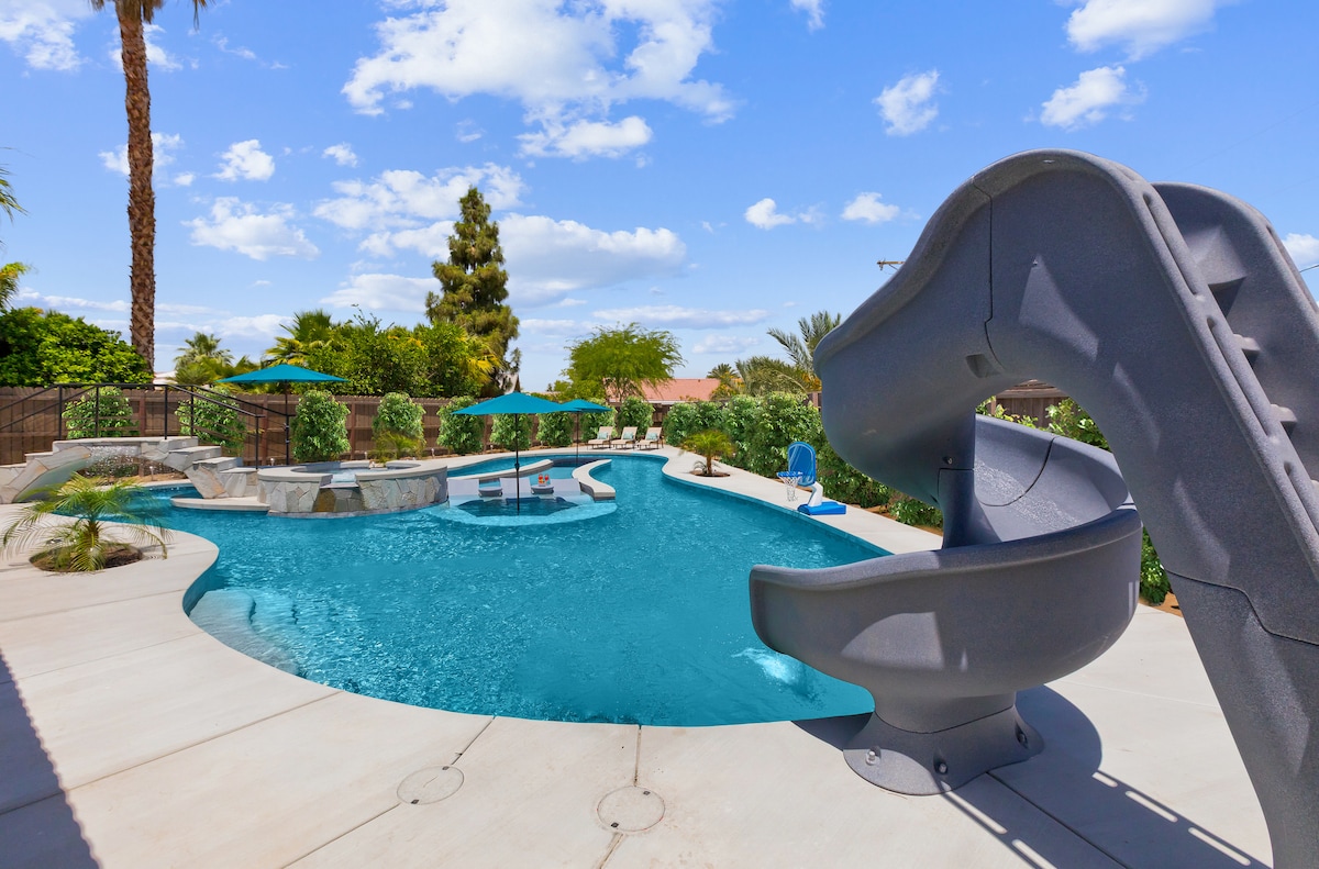 A spacious backyard features a large pool with crystal clear water, complemented by a slide and stylish loungers. Umbrellas provide shade across various seating areas. Lush greenery surrounds the pool, adding to the tranquil environment. The sky is bright with scattered clouds.