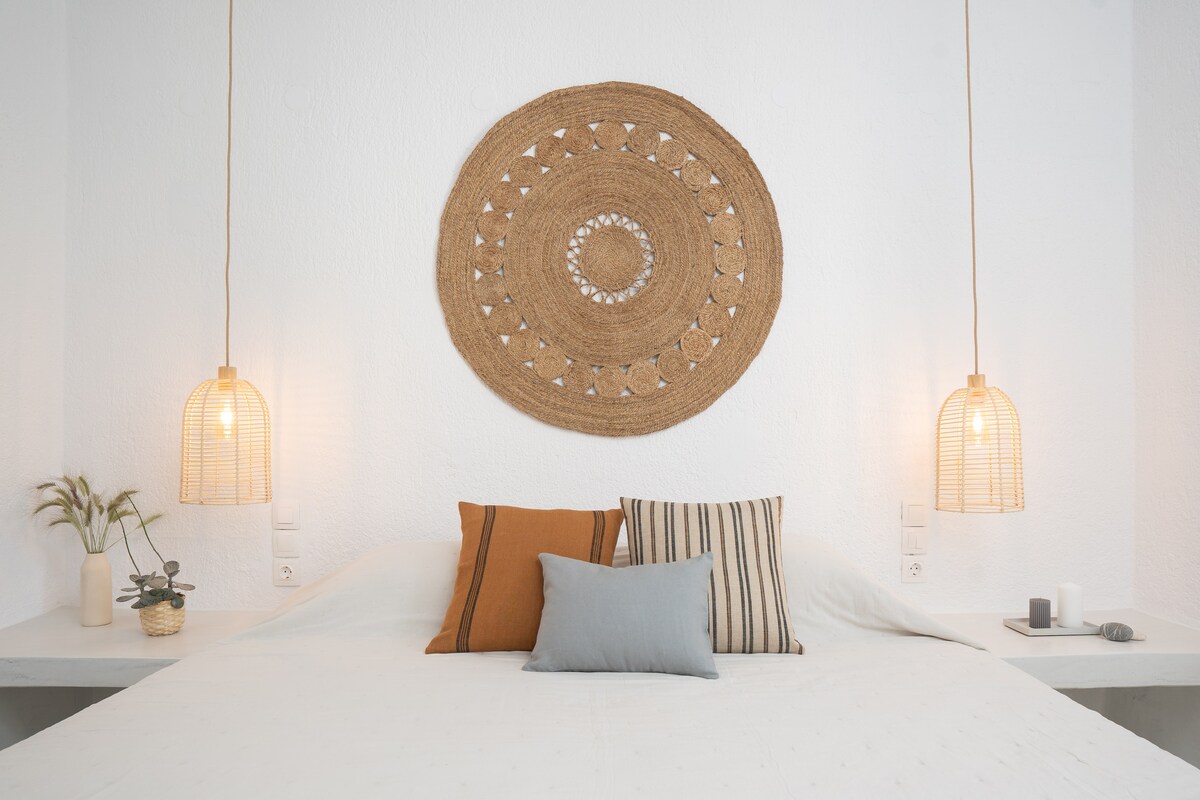 A spacious sleeping area features a neutral-colored bed adorned with decorative cushions. Natural light from woven pendant lights gently illuminates a textured wall. A minimalistic design is complemented by a decorative wall hanging and small plants arranged on bedside tables.