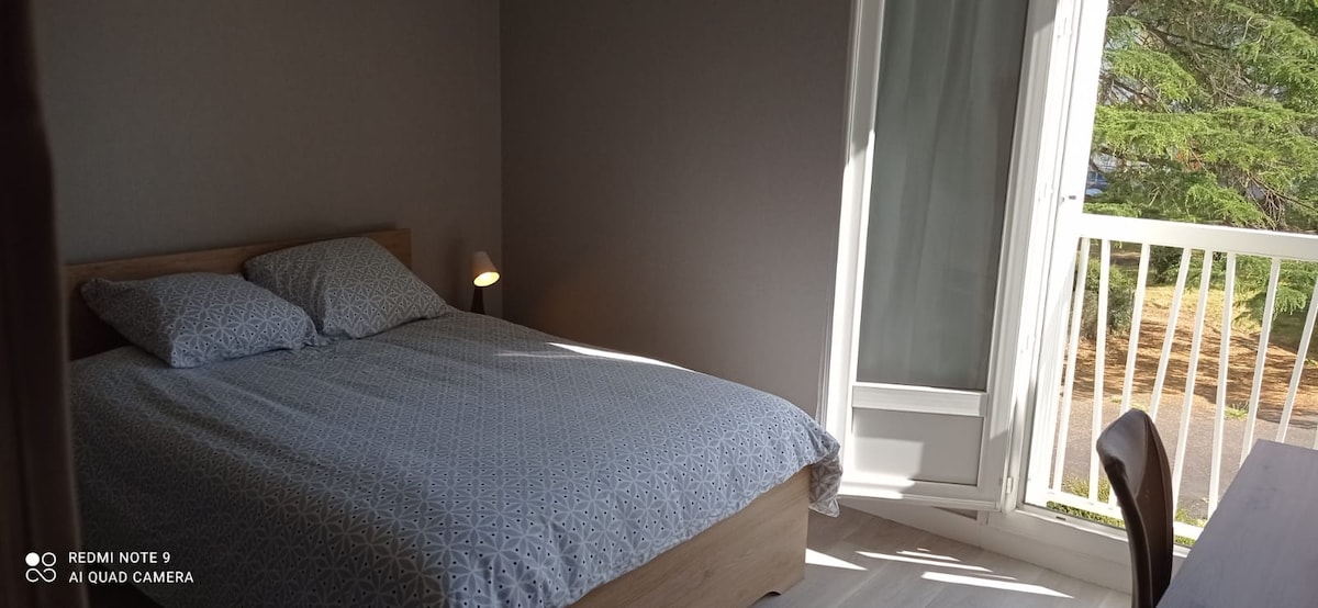 A bright bedroom features a spacious bed dressed in light bedding. A bedside lamp illuminates one side. A desk and chair are positioned nearby. A window opens to reveal greenery outside, allowing natural light to fill the room.