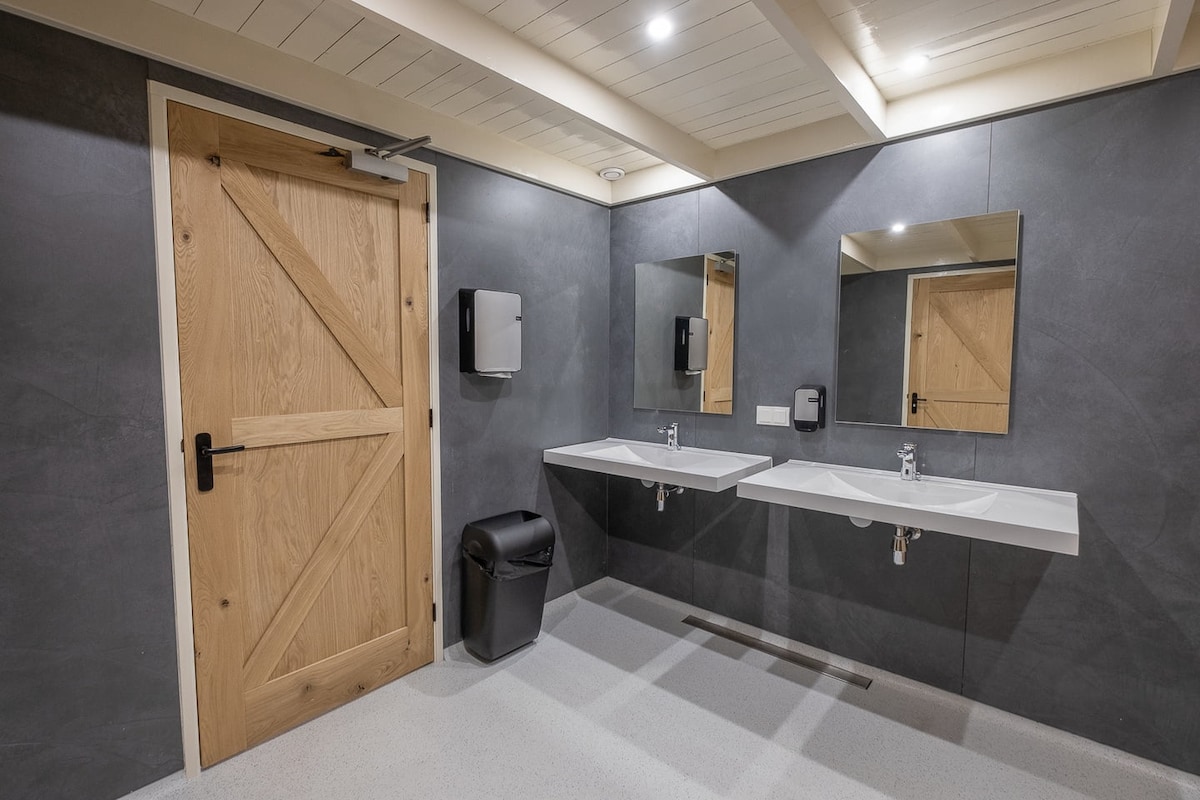 A modern restroom is equipped with two sinks and large mirrors above each. Neutral-colored walls are complemented by a warm wooden door. A waste bin is located beside the sinks, and soap dispensers are mounted on the wall for convenience.