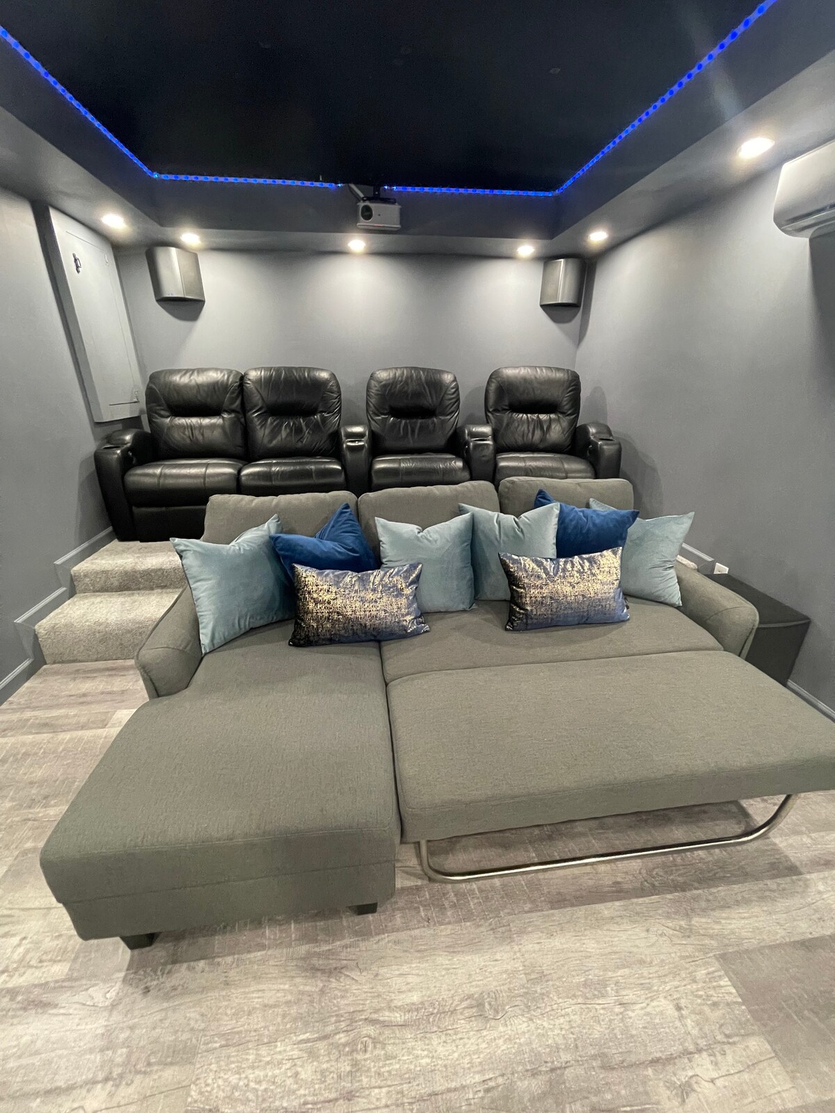 A theater room features a combination of leather reclining chairs and a spacious sleeper sofa adorned with decorative pillows in shades of blue and gold. Soft lighting and a starlit ceiling enhance the ambiance for an enjoyable movie experience.