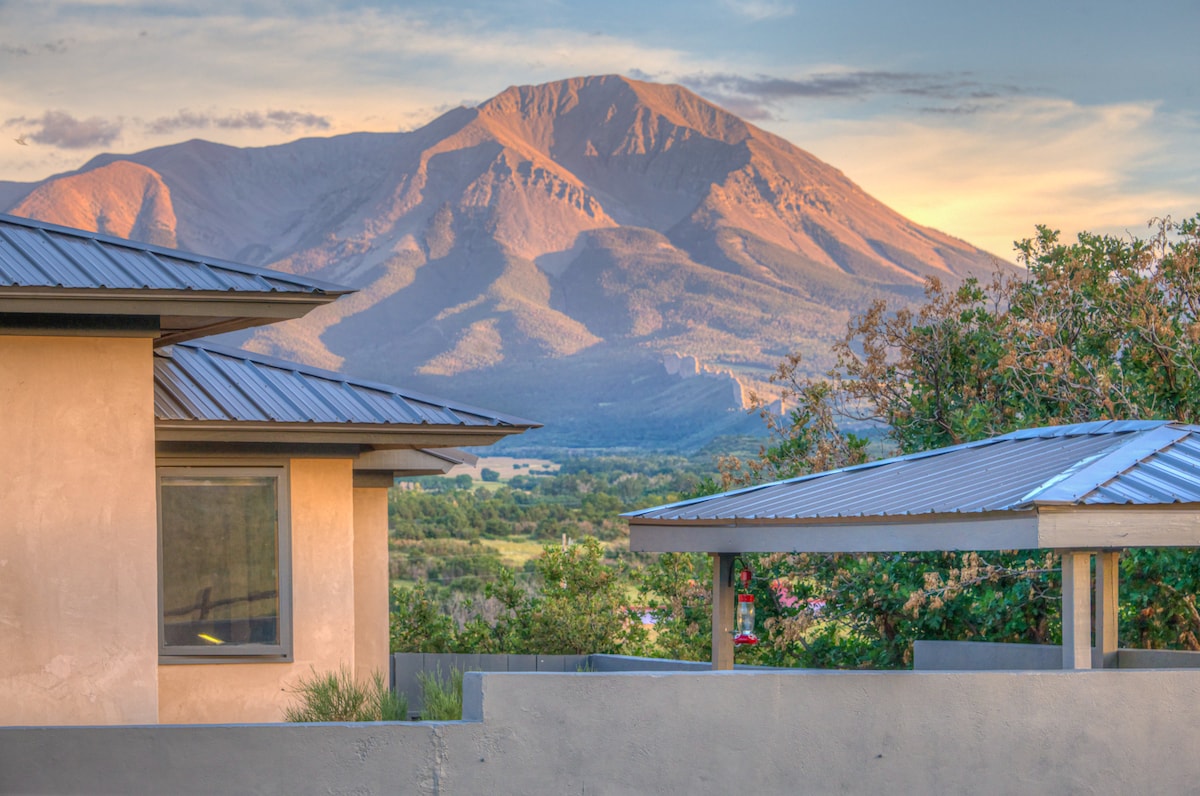 A view of the Spanish Peaks is captured, showcasing their impressive silhouette against a colorful sky. The image reveals a combination of architectural elements, including the roof of the guesthouse and a covered patio, framed by green foliage.