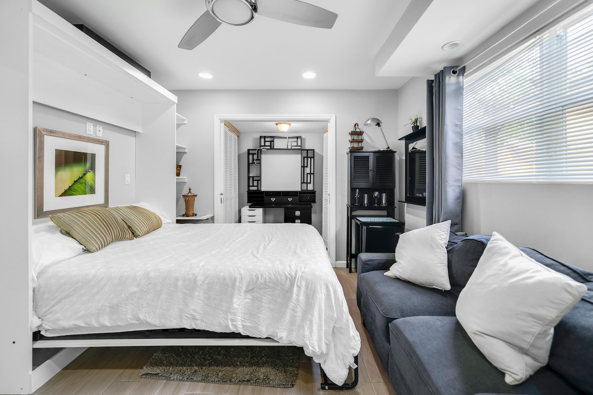 A compact studio features a Murphy bed with soft linens, set against a light wall. A cozy seating area with dark cushions sits adjacent. Natural light enters through large windows, and a stylish wall unit provides storage and display options.