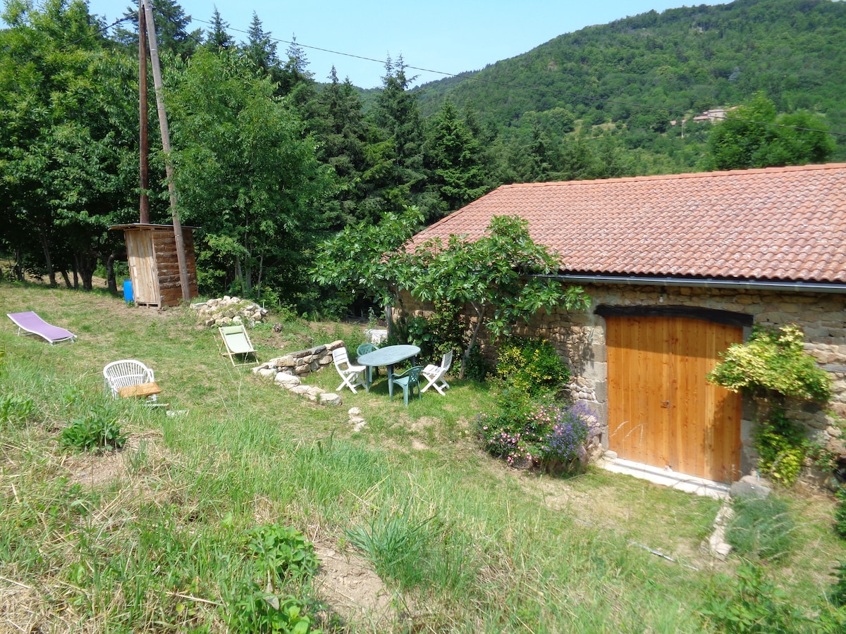 An outdoor space is shown, featuring a rustic wooden door leading into the gîte. A circular table surrounded by white chairs is visible, alongside a stone path. Sun loungers are positioned on the grassy area, bordered by trees and shrubs, creating a serene atmosphere.