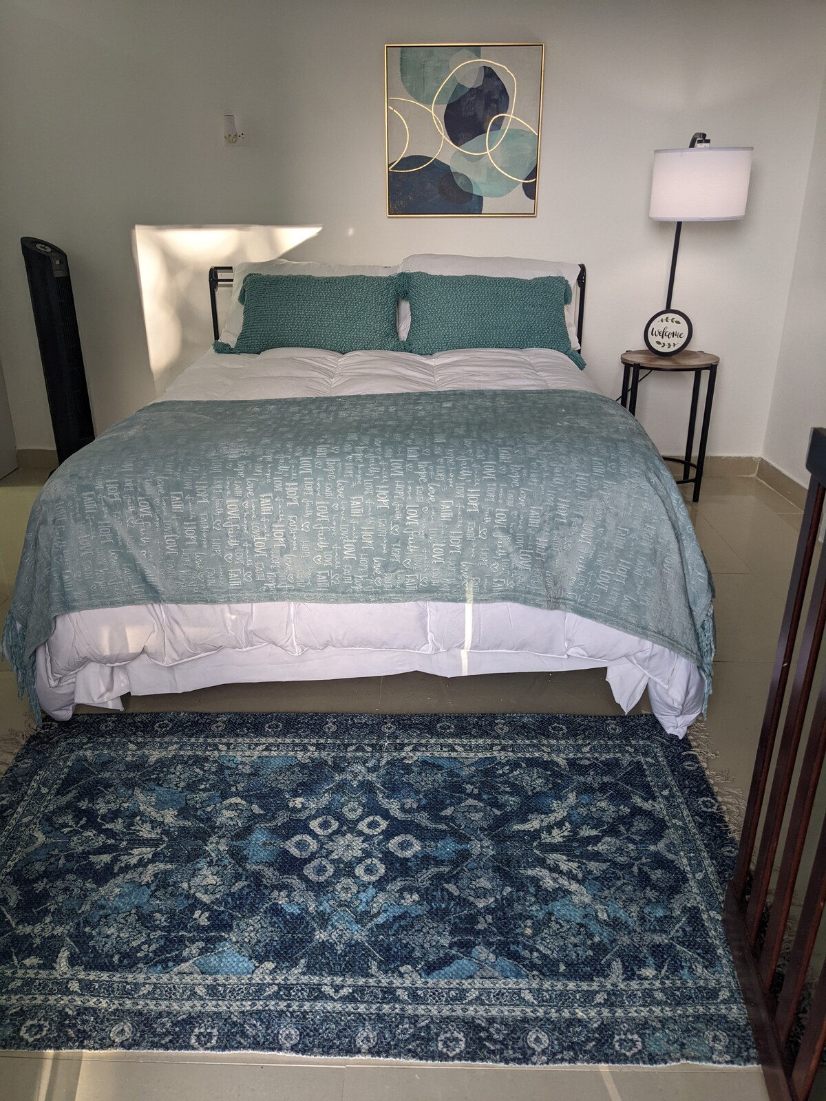 A serene sleeping area features a spacious bed with a light duvet and decorative pillows. A soft blue area rug is positioned beneath the bed, complementing the overall color scheme. A modern lamp provides gentle lighting, while a piece of abstract art adorns the wall.