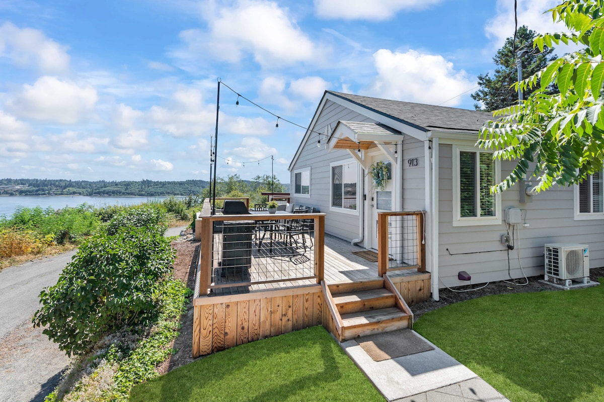 A modern exterior features a welcoming front deck, surrounded by greenery and offering views of the water. The deck is equipped with patio seating and lights, creating a comfortable outdoor space for dining and relaxation. A grassy area is visible, perfect for yard games.