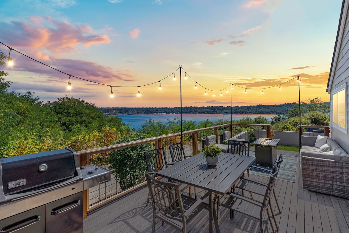 A spacious deck overlooks serene water views, adorned with string lights that cast a warm glow. A dining table is set for outdoor meals, complemented by a grill nearby. Comfortable seating is arranged for relaxation, creating an inviting space for gatherings.