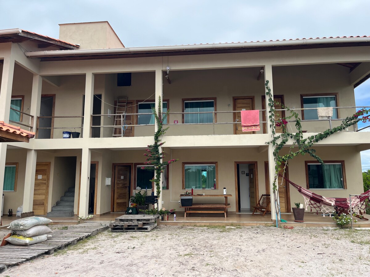 A two-story building is depicted, featuring a spacious balcony with wooden railings. The ground level has multiple doorways, and colorful fabrics are hung outside. A hammock is visible beside a wooden bench, with sandy ground and potted plants in the surrounding area.