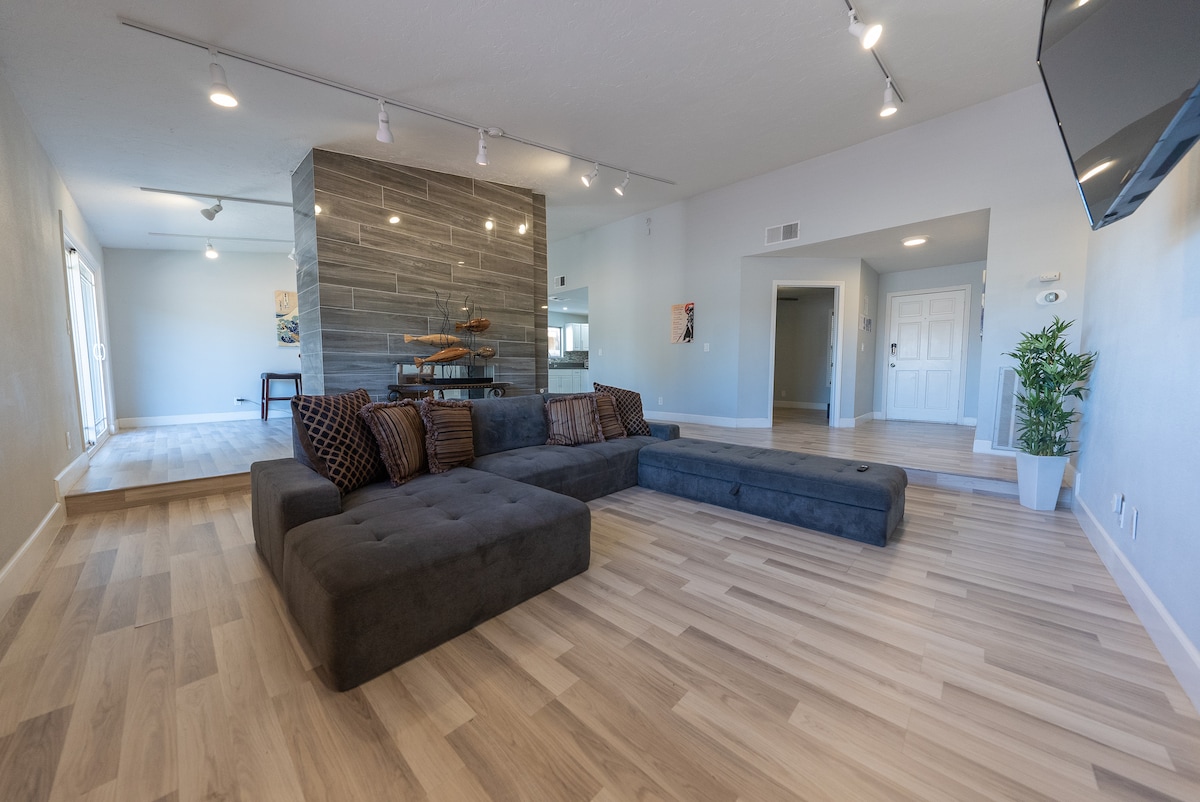 A modern living room features a large, dark sectional sofa with plush cushions. Natural light floods the space through sliding glass doors, highlighting the light wood flooring. A contemporary stone accent wall adds texture, while potted greenery enhances the inviting atmosphere.