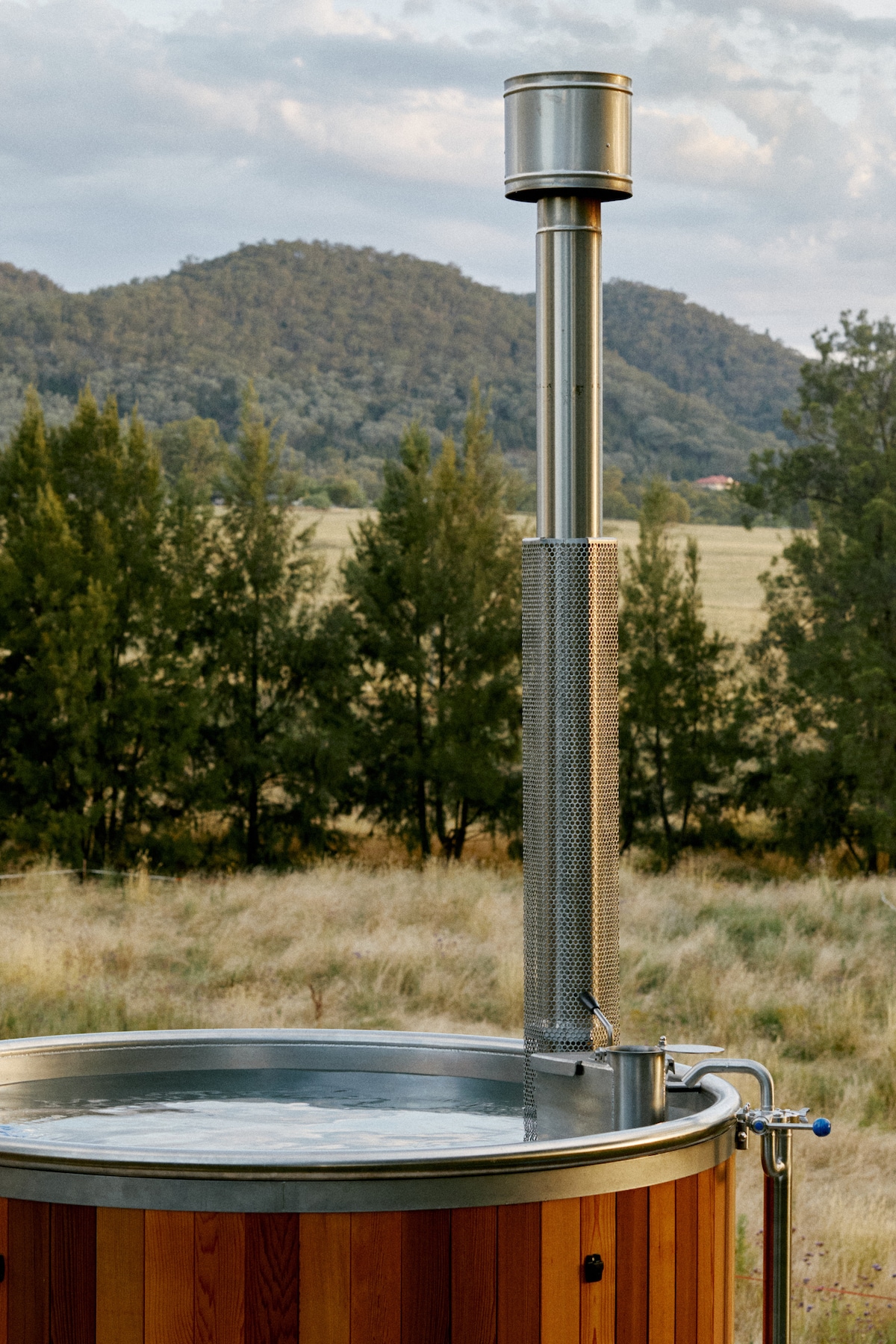 A wood-fired hot tub is displayed outdoors, featuring a stainless steel chimney and a wooden surround. The scenery includes rolling hills and a backdrop of trees, with an open field extending beyond, creating a serene and natural setting.