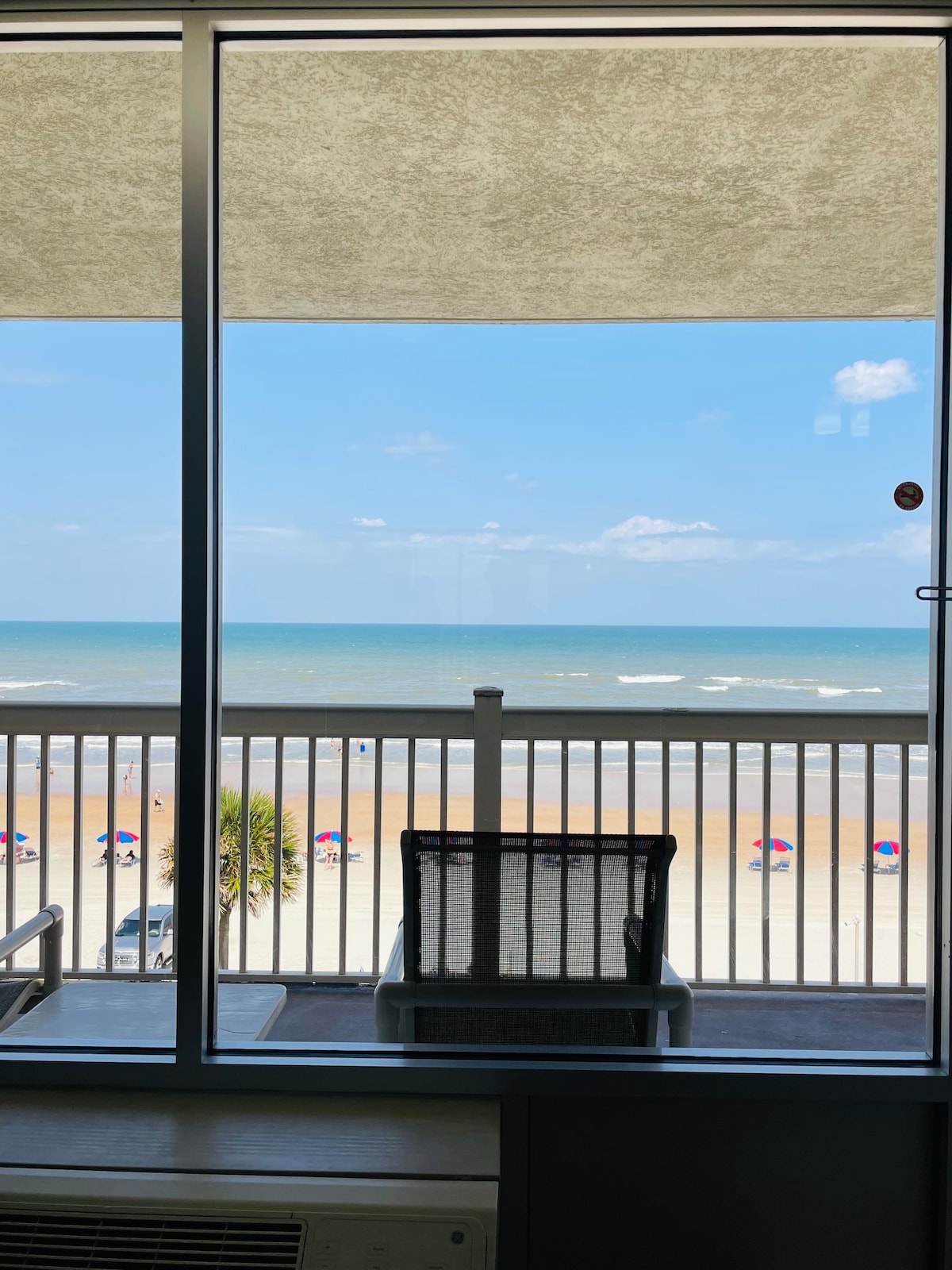 A clear view of the ocean is framed by a large window, showcasing gentle waves and sandy beaches below. Beach umbrellas in various colors add a vibrant touch to the shoreline, while a comfortable chair and small table suggest a relaxing space for enjoying the scenery.